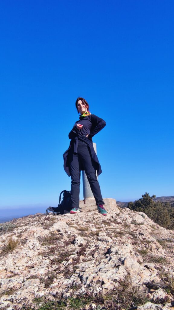 Guía de Tembo Tour posando sonriente en la cima de una montaña bajo un cielo azul intenso, con ropa técnica de senderismo.