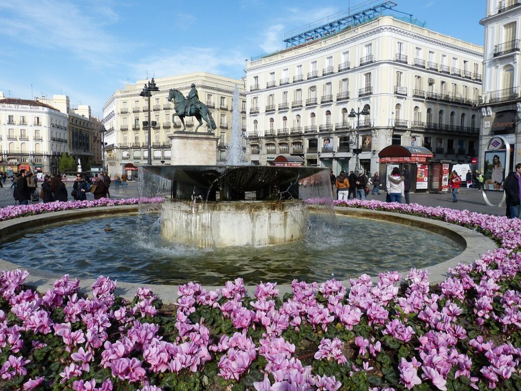 Fuente rodeada de flores en la Puerta del Sol de Madrid, con la estatua ecuestre de Carlos III al fondo.